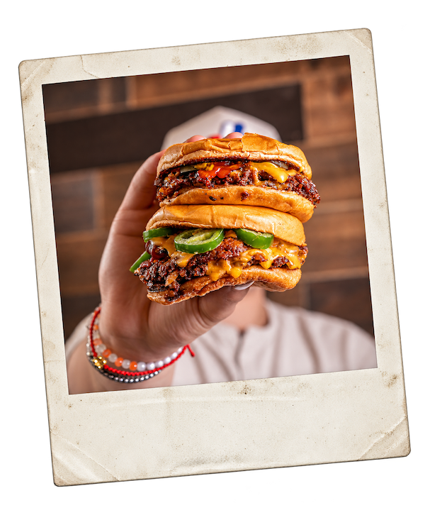 A person holding up a stack of two Crack Burger smashburgers topped with melted cheese and jalapeños, printed as a retro polaroid